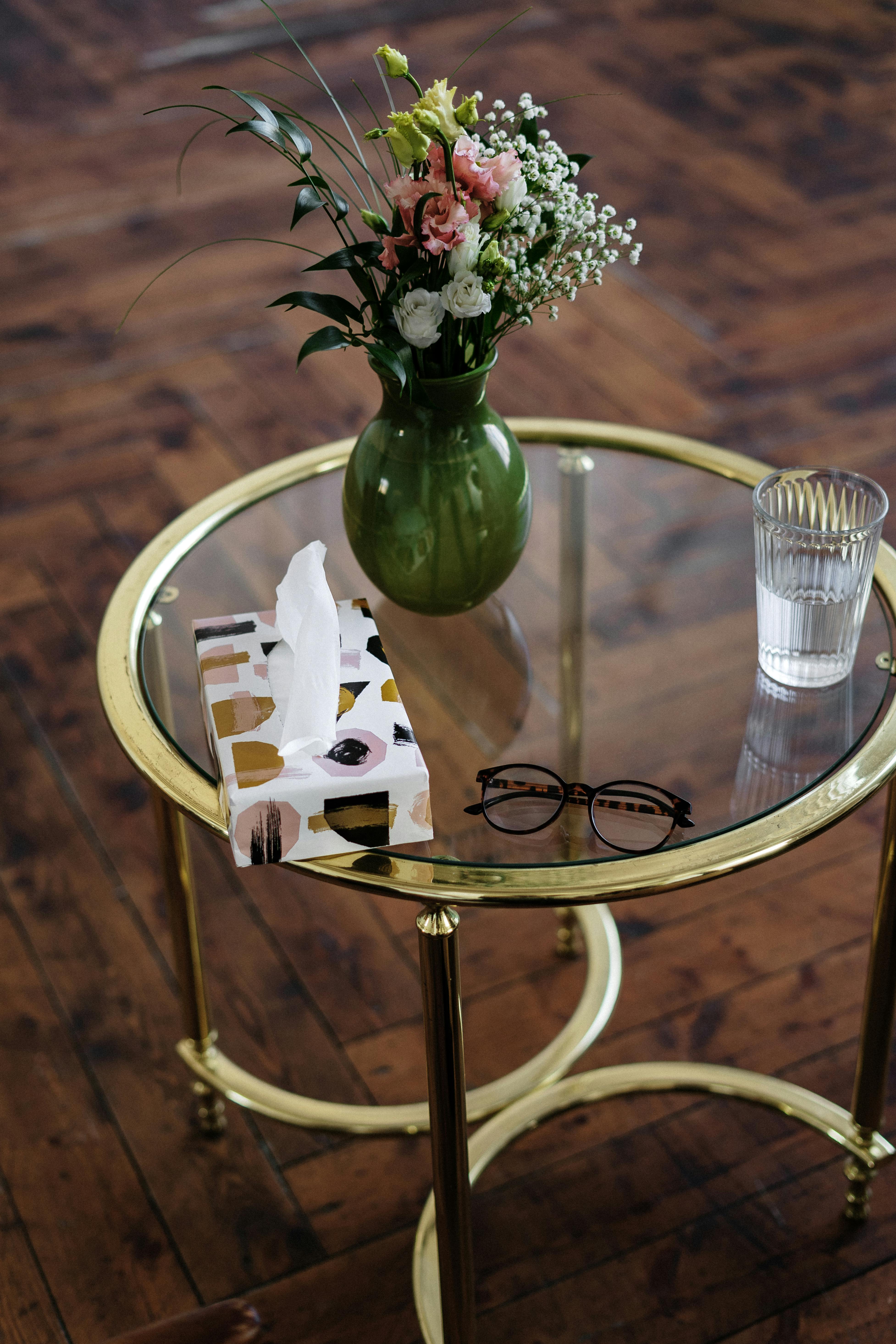 A stylish glass table with a green vase of flowers, tissue box, and reading glasses on parquet flooring.