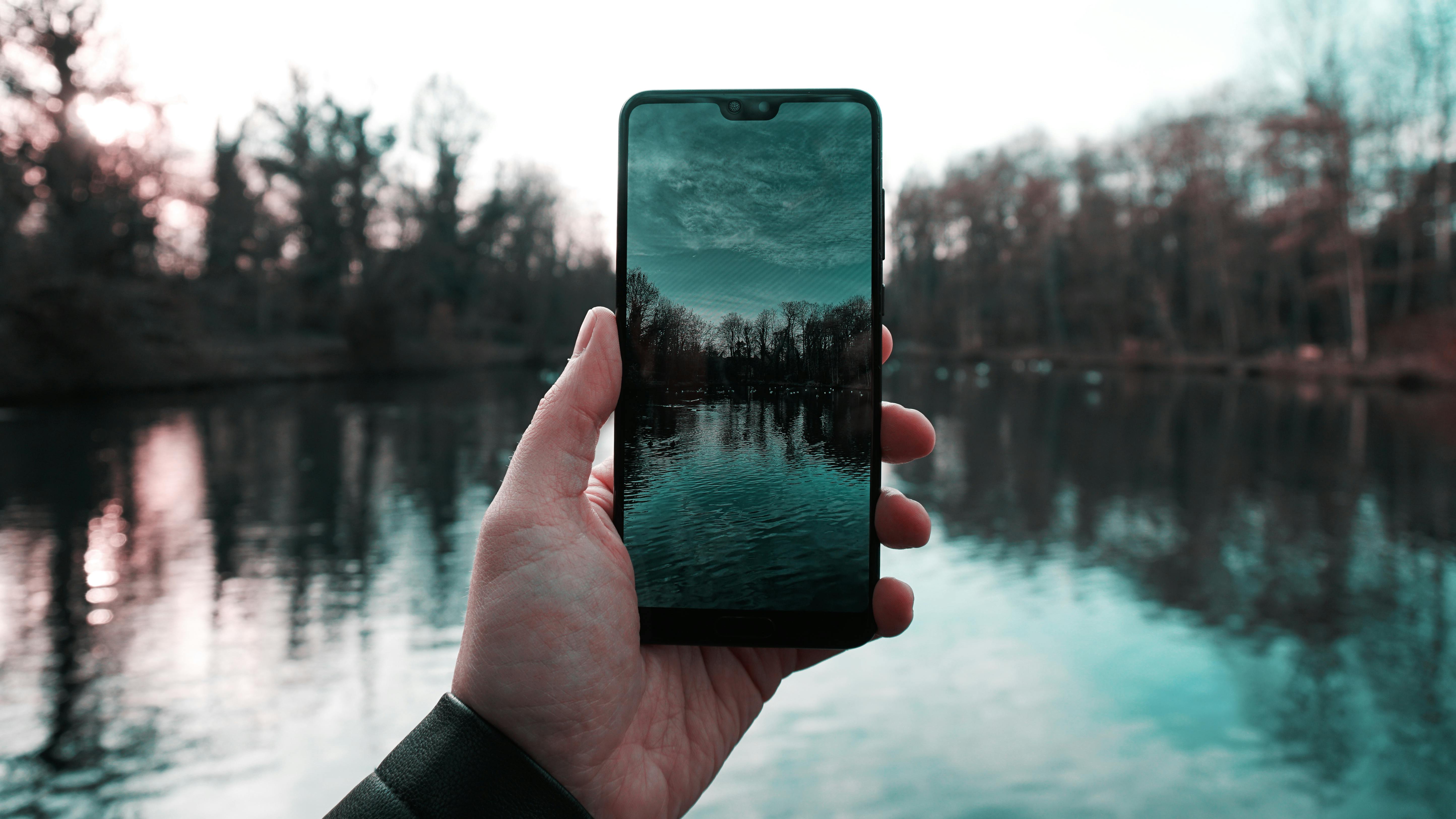 A smartphone captures a reflection of a serene lake and trees during daytime, blending technology and nature.