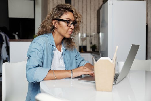 A woman in a denim jacket working remotely from home on her laptop, enjoying a casual workspace.