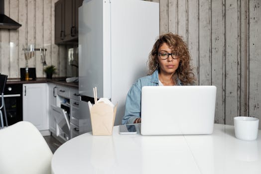 A woman using a laptop at a kitchen table, surrounded by modern appliances.