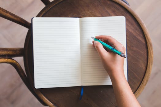 Close-up of a hand writing in a notebook with a pen on a wooden chair.