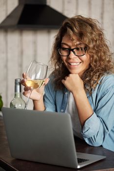 一位戴眼镜的女士面带微笑，手持酒杯，在室内用笔记本电脑工作