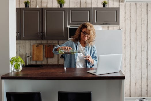 Woman pours wine while working on a laptop in her modern kitchen at home.