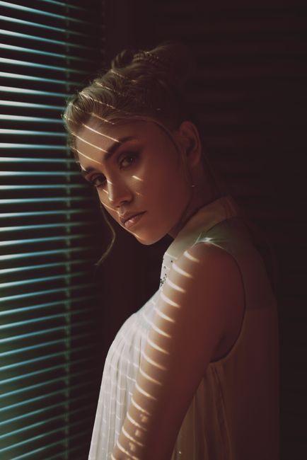 Stylish female portrait with dramatic lighting through window blinds, creating a moody atmosphere.