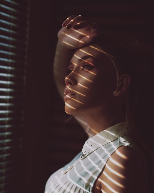 A woman gazes through window blinds, bathed in dramatic shadows and light, conveying introspection.
