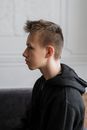 一个十几岁、穿着黑色连帽衫的男孩坐在室内，侧身肖像中露出若有所思的表情