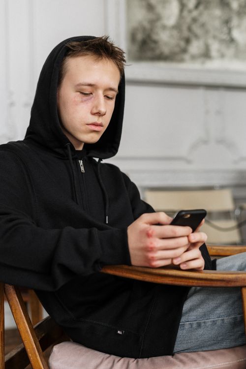 Free Teen boy in hoodie with black eye using smartphone indoors, focused expression. Stock Photo