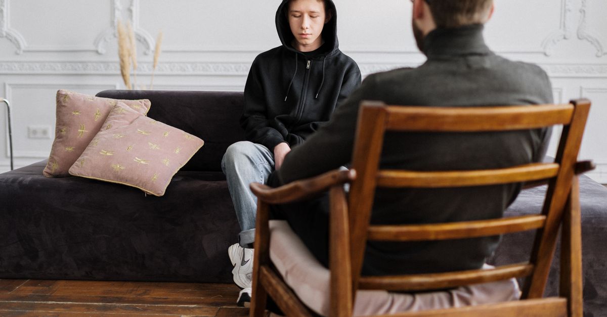 A teenager in hooded sweatshirt talks with a therapist in a modern, minimalistic studio.