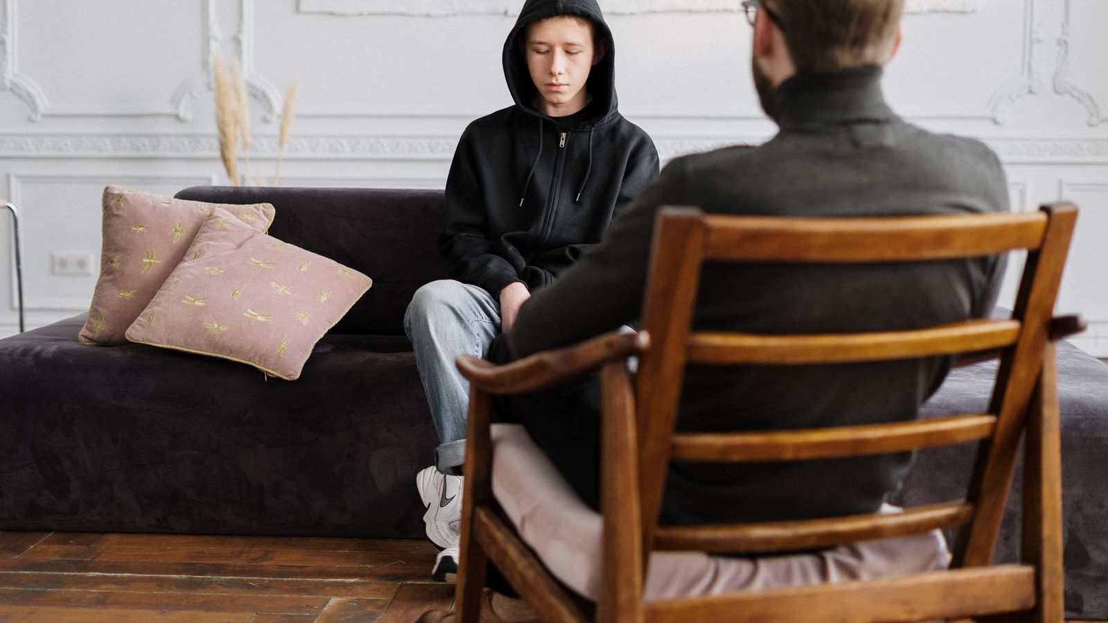 A teenager in hooded sweatshirt talks with a therapist in a modern, minimalistic studio.