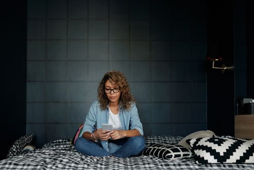 一名戴眼镜的女子坐在卧室舒适的床上，用着智能手机
