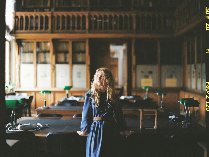 Blonde woman in blue dress standing in a vintage library with wooden interiors and soft lighting.