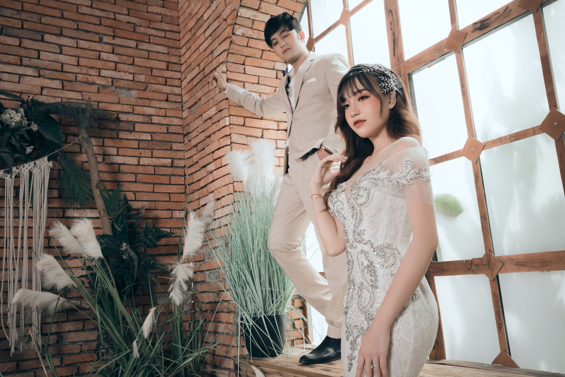 Pensive Asian well dressed groom leaning hand to wall and looking at charming slender fiance with makeup in elegant white bridal gown looking away in cozy room decorated with plants