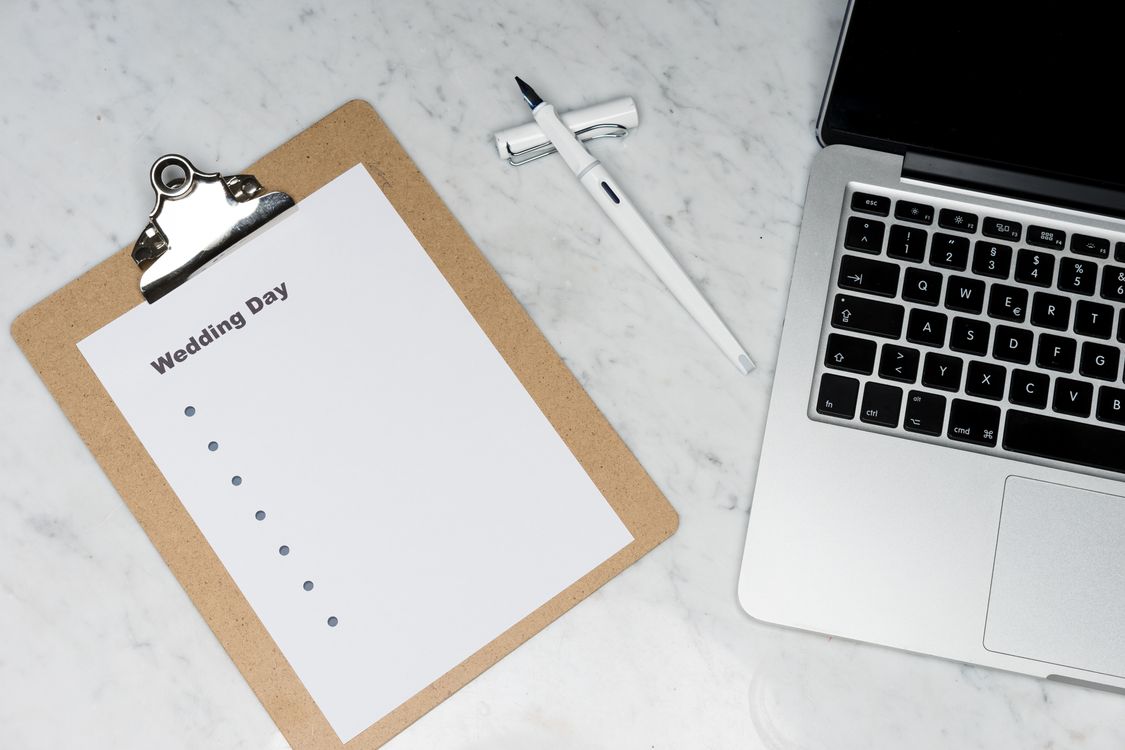 A wedding planning desk with a clipboard, laptop, pen and notes laid out on a light surface
