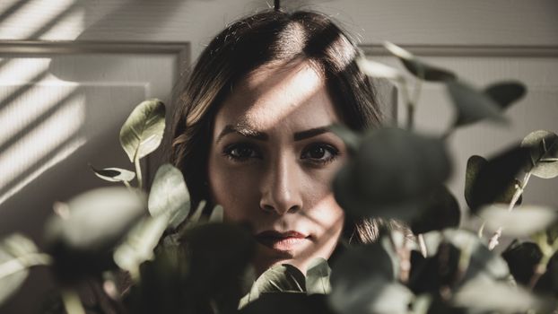 A close-up portrait of a woman partially hidden by green leaves, with dramatic light and shadow play.