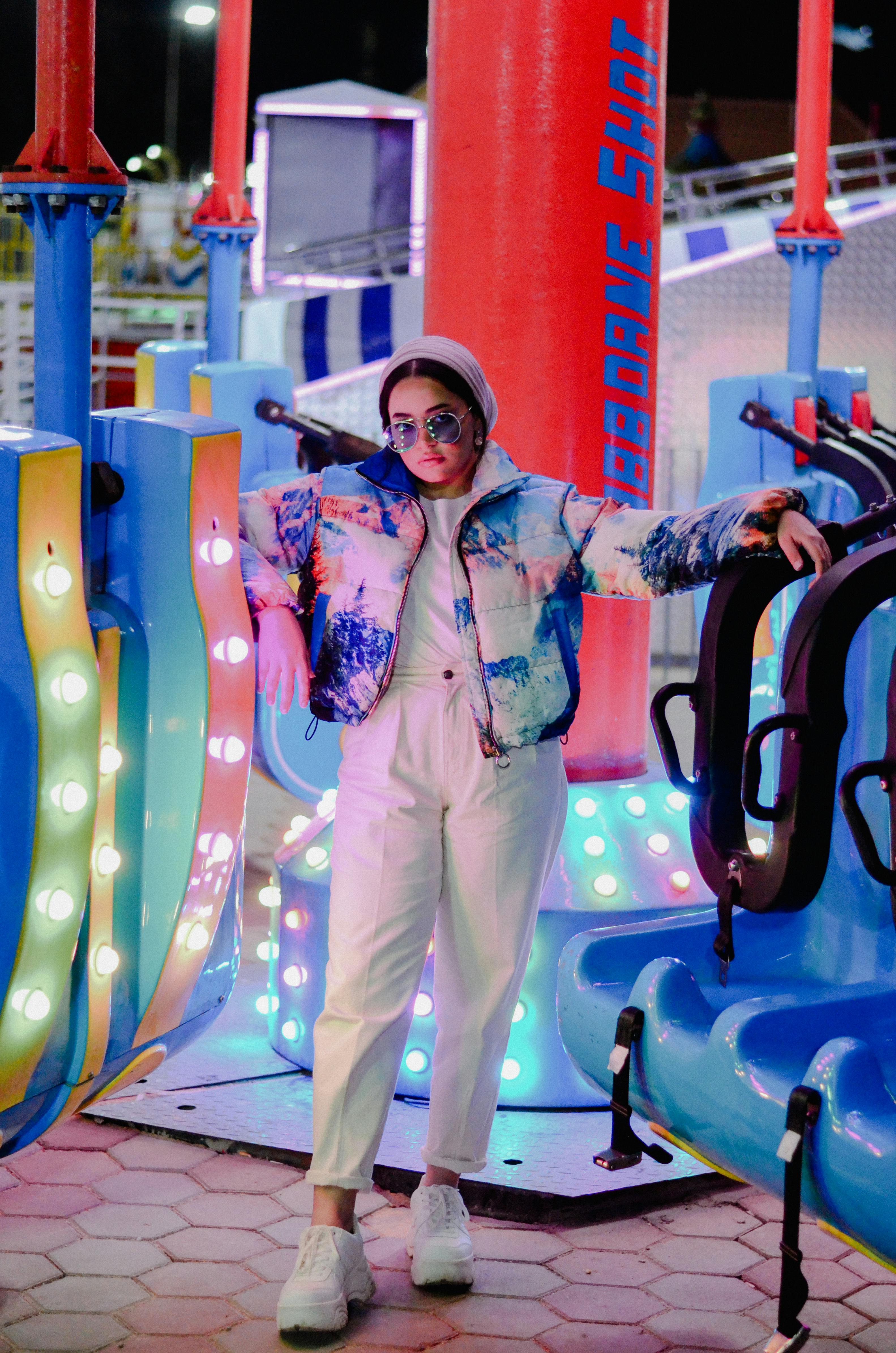 Stylish woman in trendy outfit posing at a colorful amusement park at night.