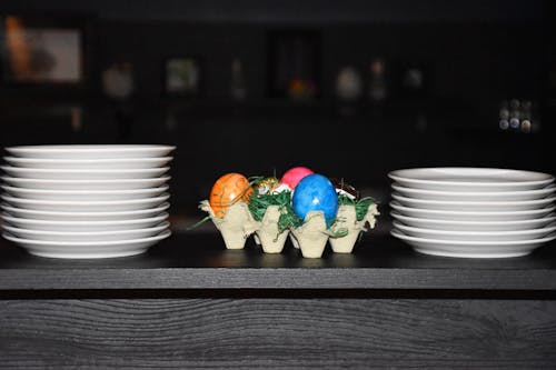 Multi colored Easter egg on carton between bunches of white plates on black wooden table in bright room