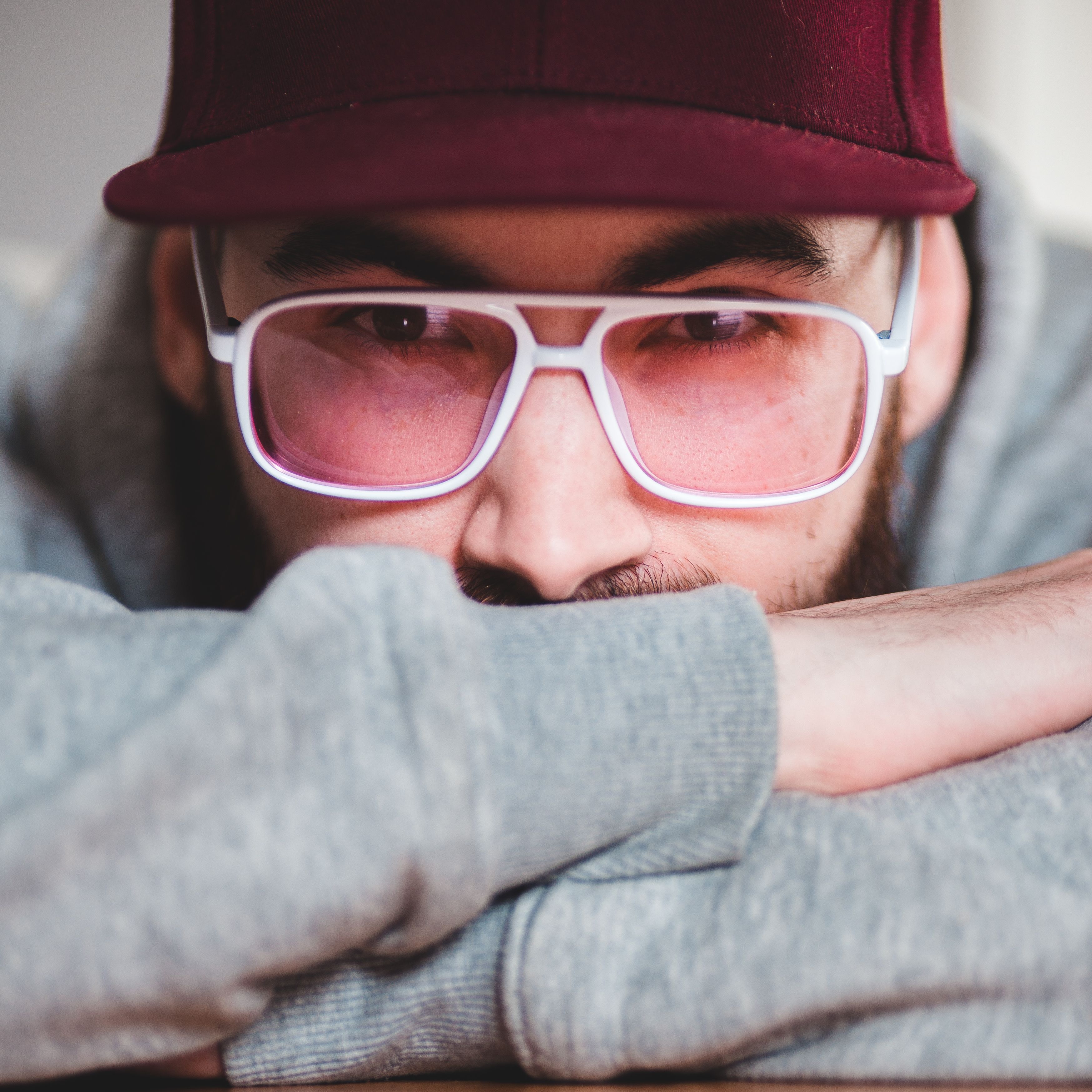 A young man wearing a modern look with pink-tinted glasses, cap, and casual attire, gazing thoughtfully.