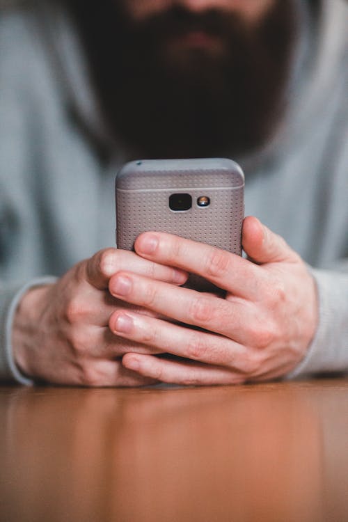 Soft focus of crop man with beard in warm grey clothing browsing smartphone