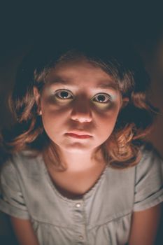 灯光下，一个卷发、穿着灰色连衣裙的无情女孩在黑暗的房间里看着镜头