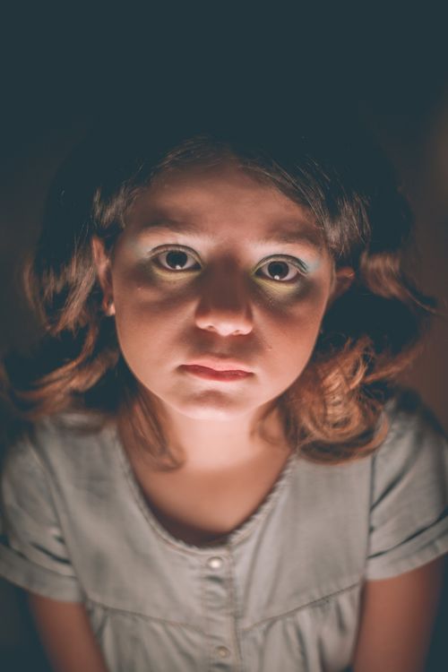 Free Emotionless girl with wavy hair in grey dress in light of lamp looking at camera in dark room Stock Photo