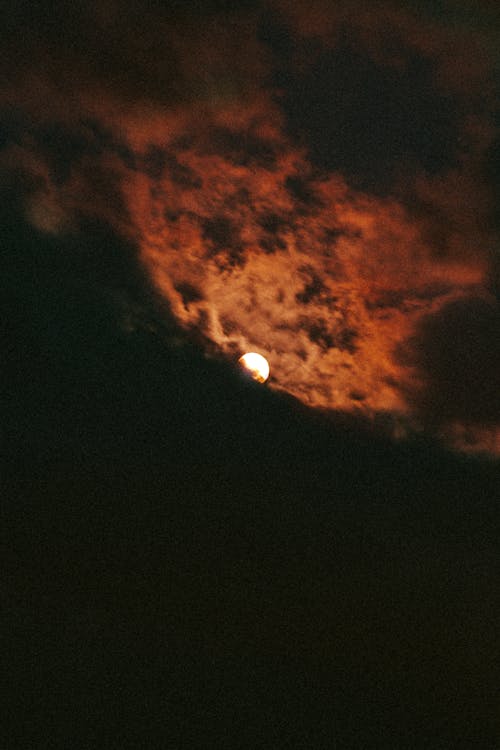 Dramatic bright glowing moon in black sky surrounded by orange clouds late at night