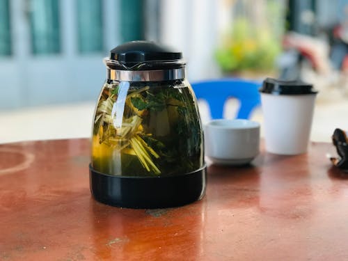 Free Transparent shining teapot with green tea and fresh mint leaves with thin stalks on wooden table near ceramic cup and disposable glass of coffee in street cafeteria in morning Stock Photo