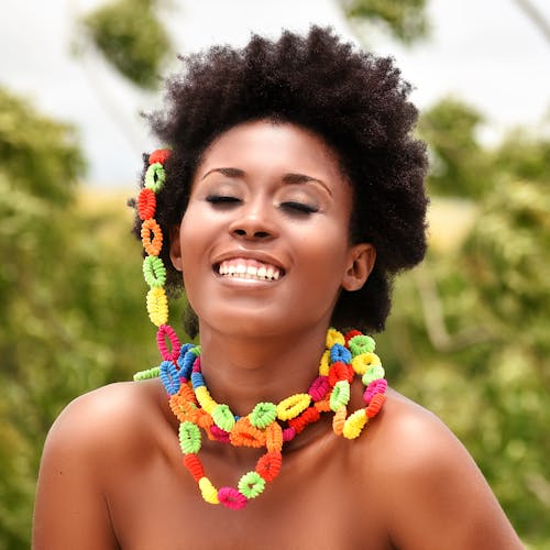 Young smiling African American naked female with Afro hairstyle and colorful beading of hair coils standing with closed eyes behind green trees in summer in sunlight