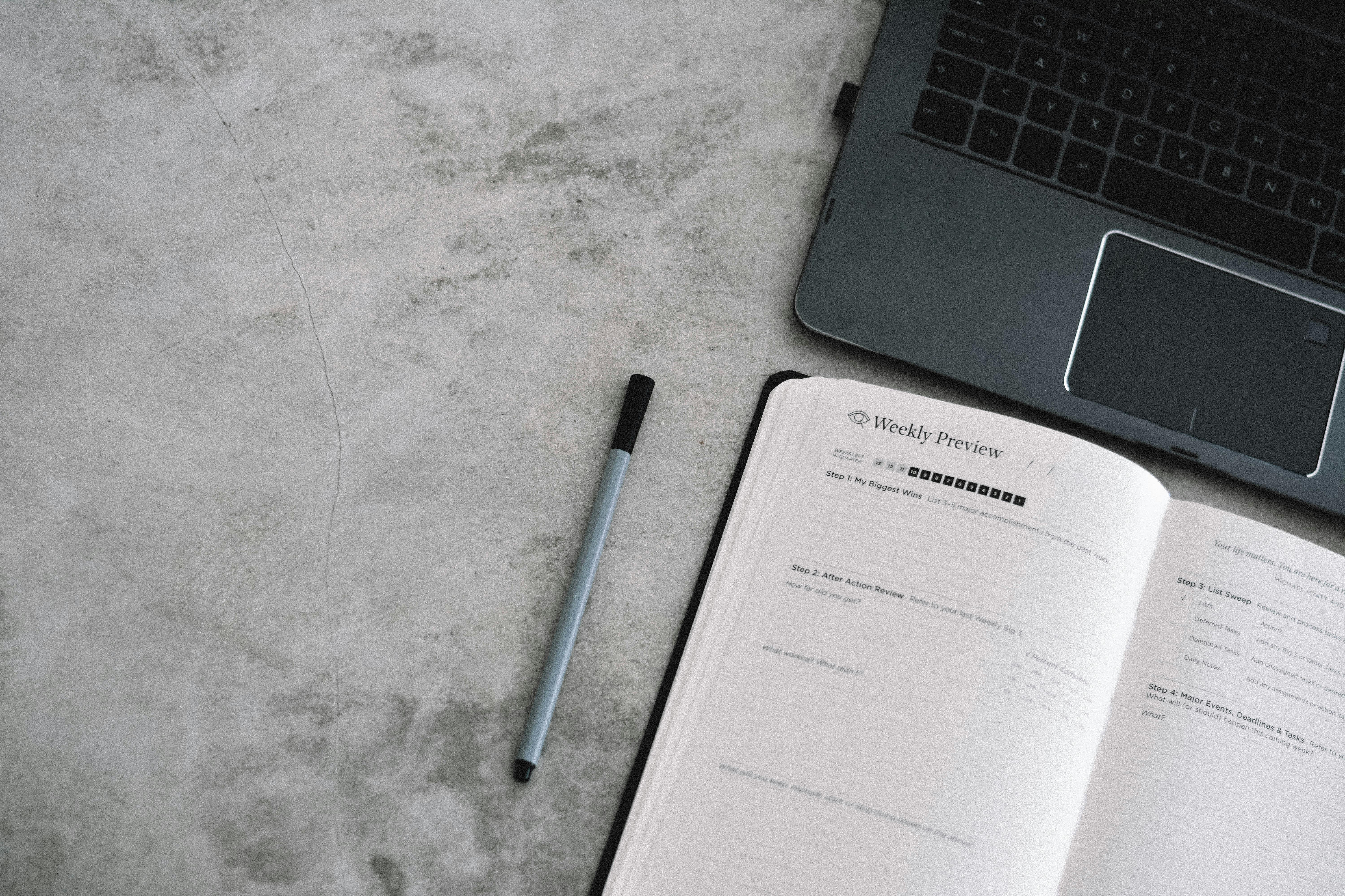 A top view of a minimalist workspace with a planner, pen, and laptop on a gray surface.