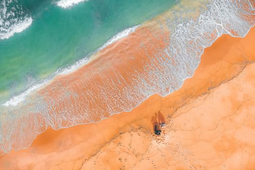 Aerial view of turquoise wavy sea with rippled water on bright brown sandy shore with anonymous couple of tourists in summertime