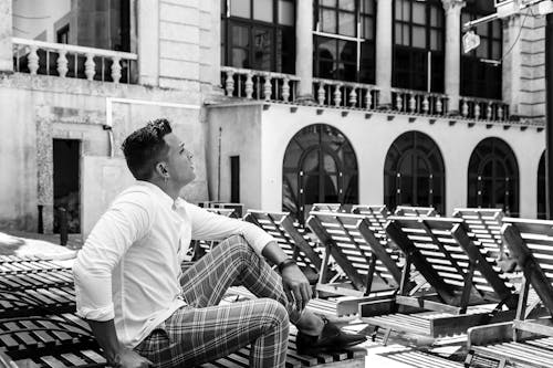 Black and white side view of fit contemplative male in stylish outfit with creative haircut looking away while sitting on sunbed near aged house with decorative fence and arched doors