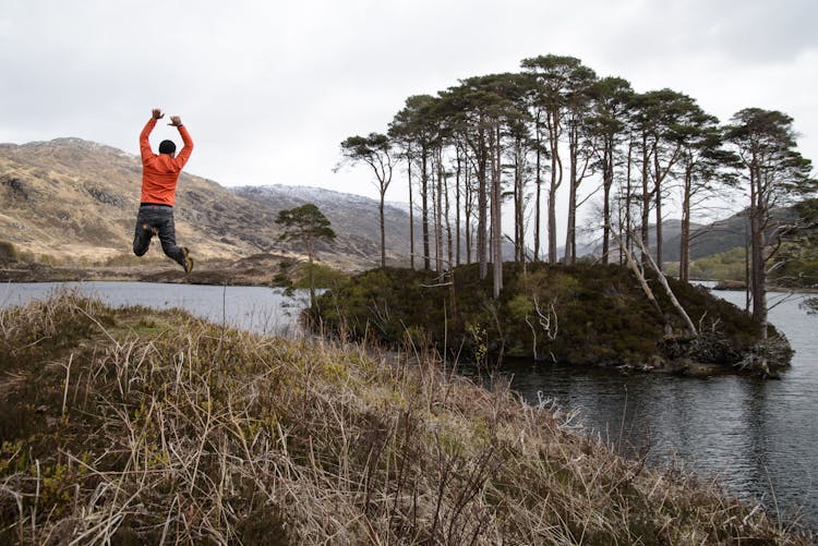 Man In Orange Long-sleeved Shirt Jumping On Lake Near Tall Trees At Daytime