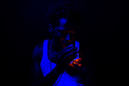 A man in a tank top lights a cigarette under moody ultraviolet lighting in a dark setting.
