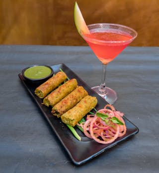 From above of yummy deep fried Vietnamese spring rolls on plate with onion slices and spicy green salsa near glass of 
cocktail in cafeteria