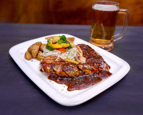 From above of tasty grilled pork steak with barbecue sauce served with salad and fried potato on table near mug of beer