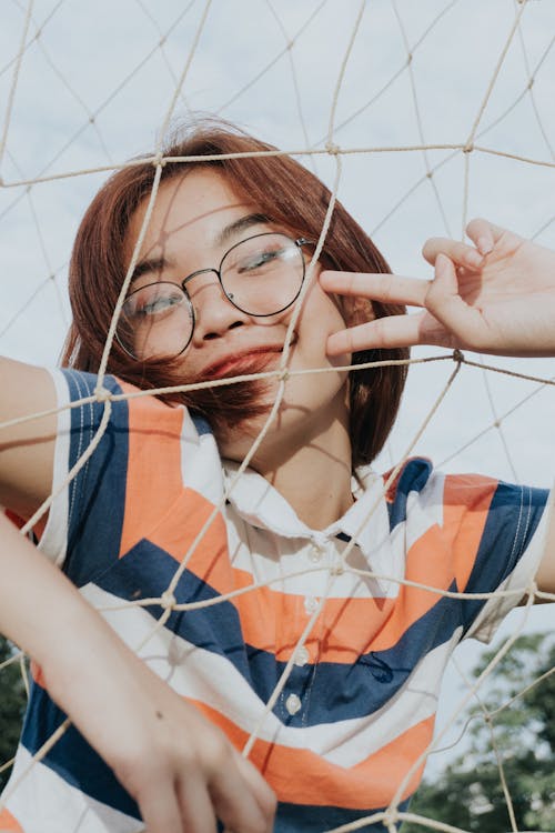 From below of slim cheerful young ethnic female in striped t shirt and eyeglasses demonstrating victory gesture while standing near grid fence in daylight and looking at camera