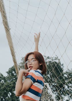 From below side view of young seductive Asian lady in eyeglasses and striped t shirt touching lips while standing near soccer goal behind trees under sky and looking at camera