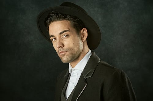 Free Portrait of stylish positive young Hispanic male wearing elegant black suit and hat standing against black background in studio and looking at camera Stock Photo