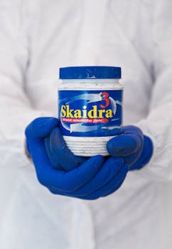 Photo by Elīna Arāja Hands wearing gloves holding a blue jar of cleaning paste indoors for sanitation.