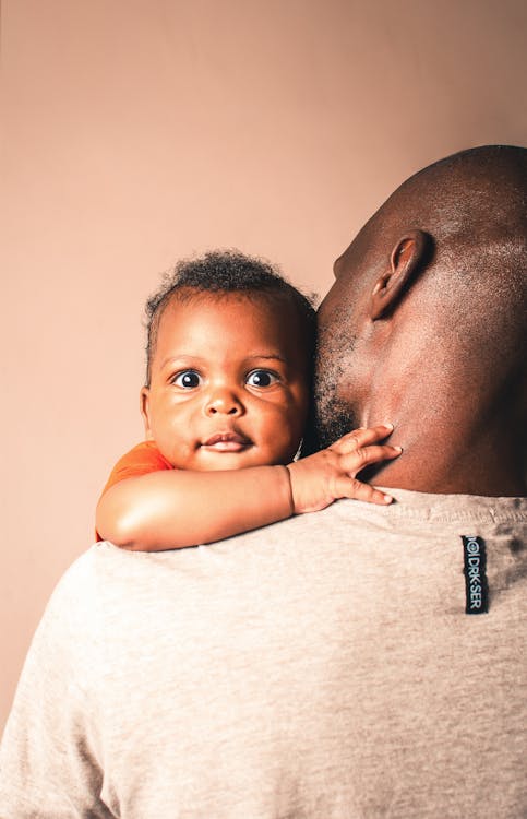 Careful African American father wearing casual gray shirt gently hugging adorable little baby and kissing cheek while standing against light pink wall