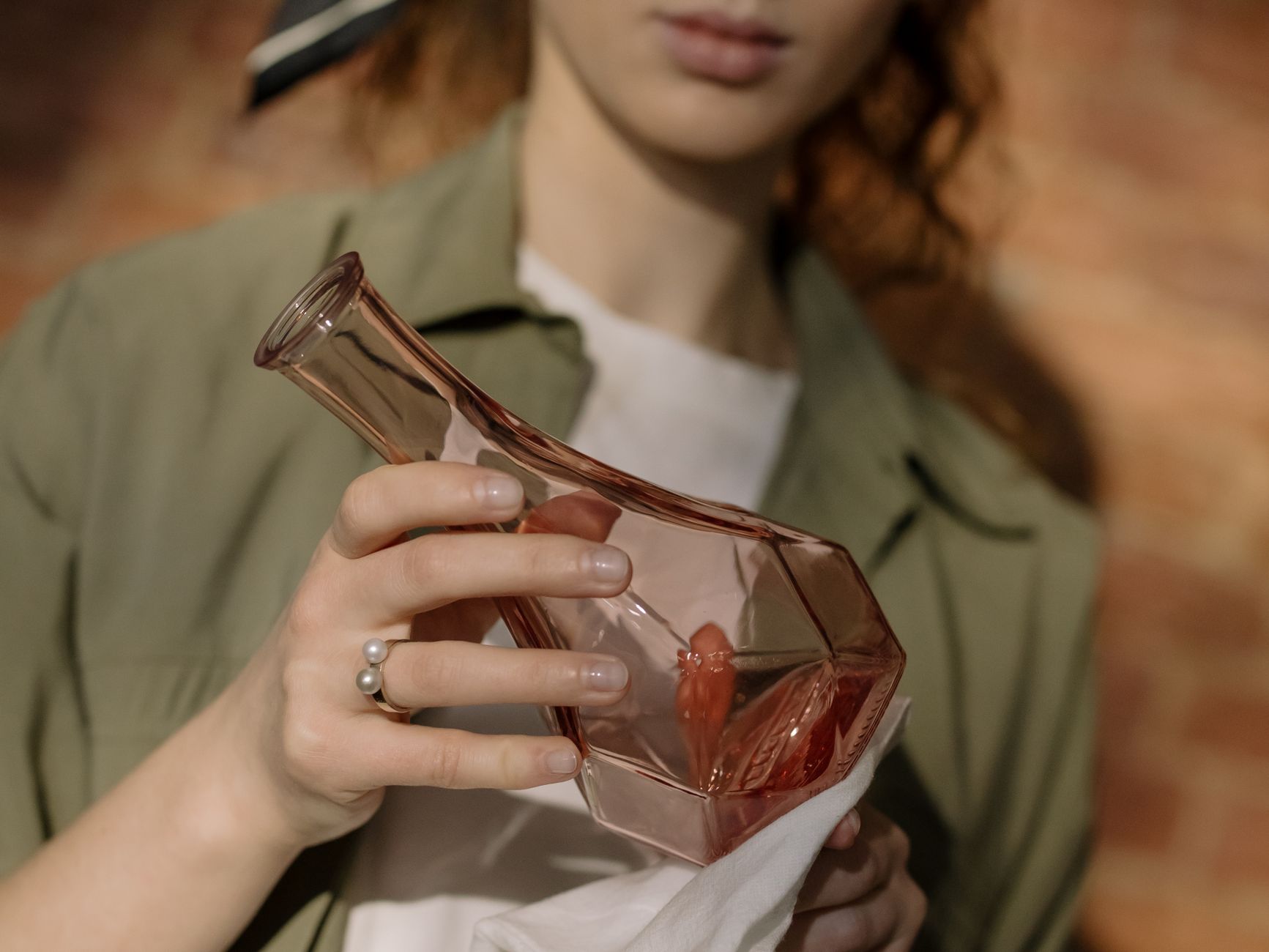 A woman carefully cleans a glass vase with a cloth beside a brick wall.