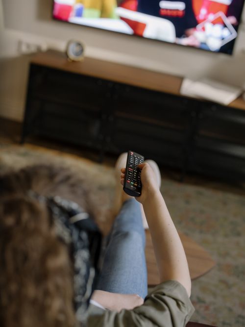 Free A woman relaxes on a couch inside her cozy home, watching television with a remote in hand. Stock Photo