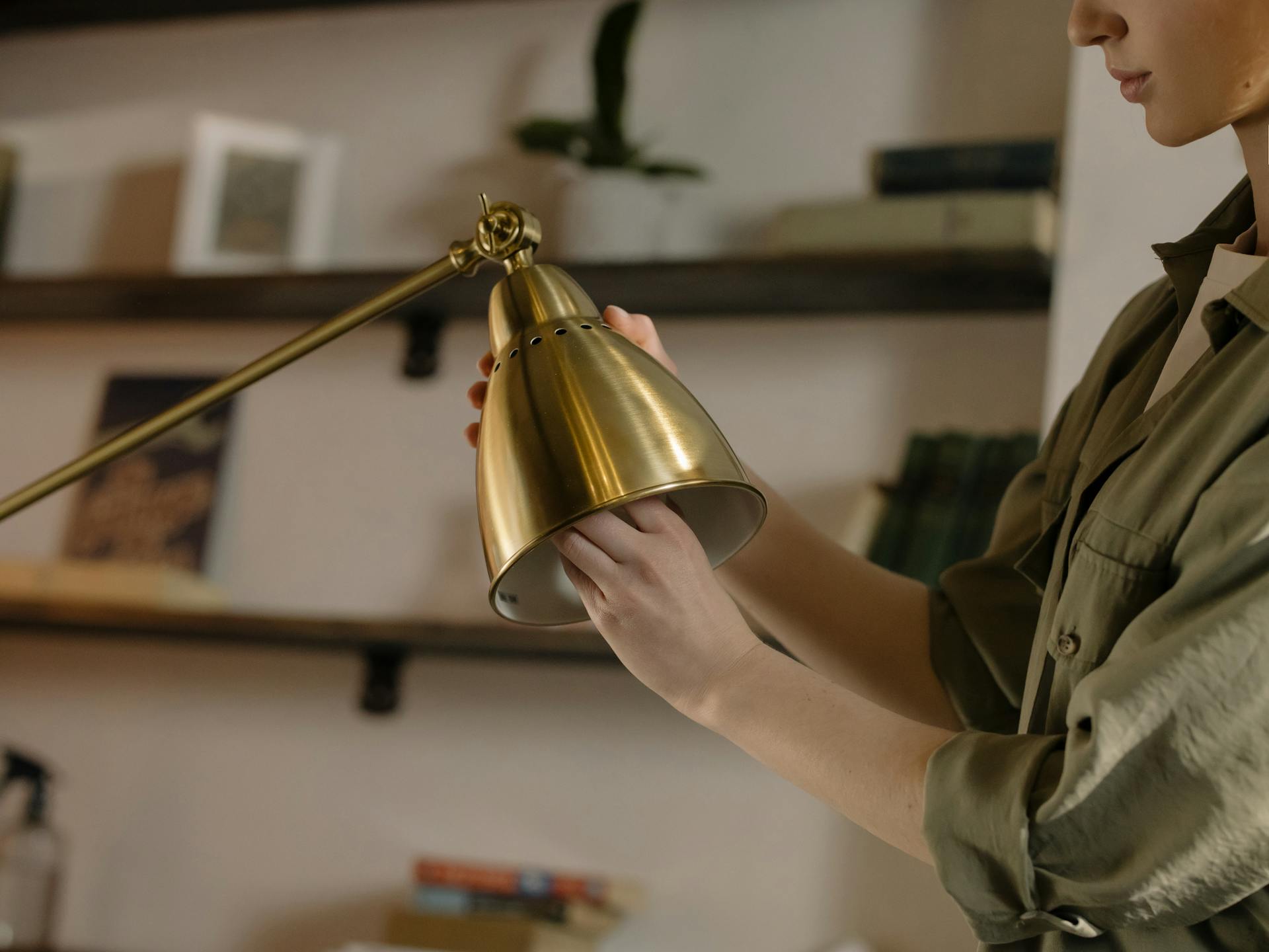 A person adjusting a brass desk lamp in a cozy home interior with shelves.