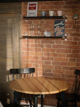 Warm kitchen corner with brick wall, round table, and shelves holding decor items and glassware.