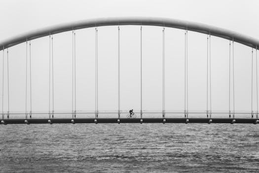 这张黑白照片捕捉到一名孤独的骑行者，在平静海面上的极简主义吊桥上骑行