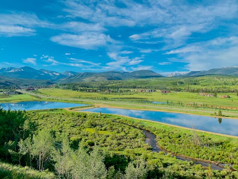 Explore a breathtaking lush green valley with a clear blue sky and distant mountains reflecting in serene water.