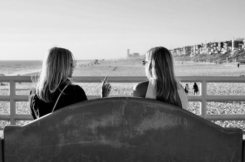 Free Back view of black and white anonymous young female friends resting on bench near sandy beach and speaking with each other on sunny day Stock Photo