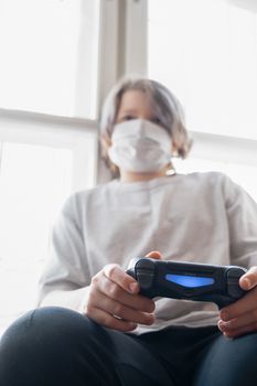 Teen sitting indoors, wearing a face mask, playing video games for leisure during quarantine.