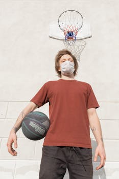 Young man with a face mask playing basketball on an outdoor court during the day.