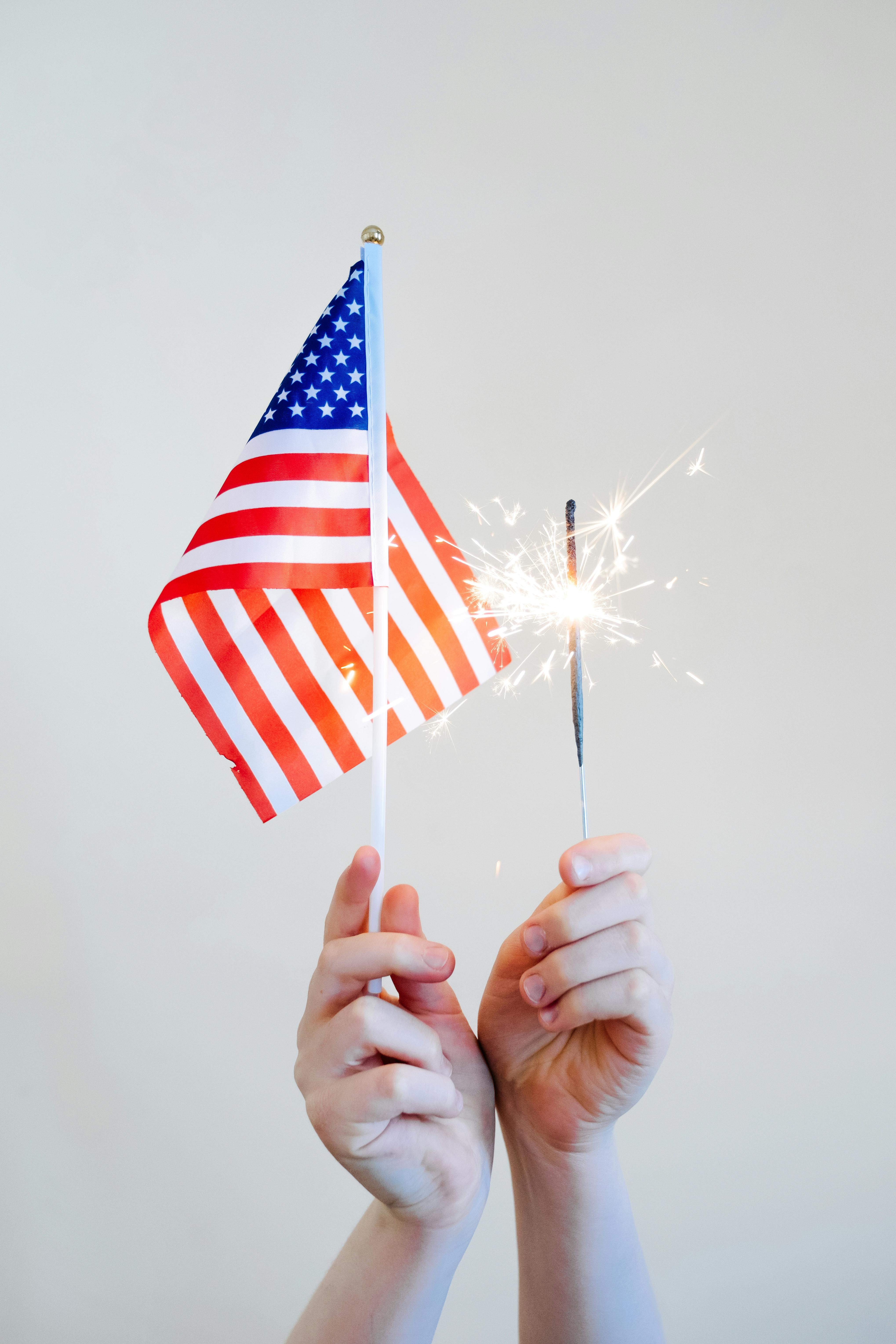 Hands holding an American flag and a lit sparkler against a white background.