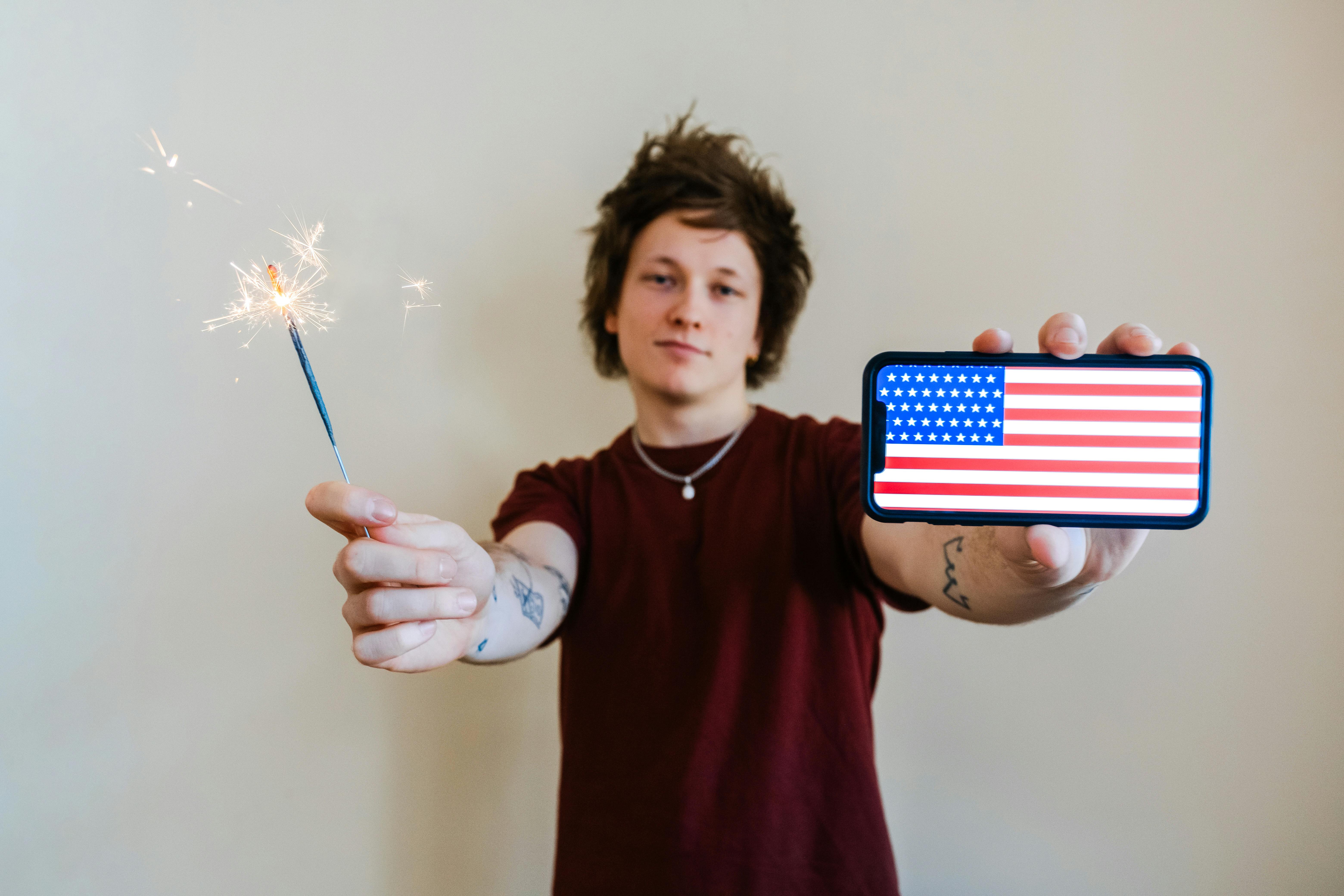 Young adult holding sparklers and a smartphone with USA flag. Celebrating with excitement.